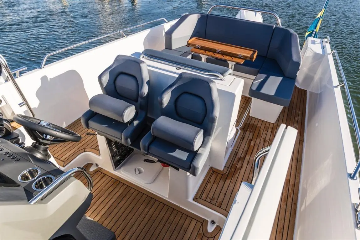 Interior of a boat featuring two seats, a steering wheel, and a wooden table on a teak deck by the water.